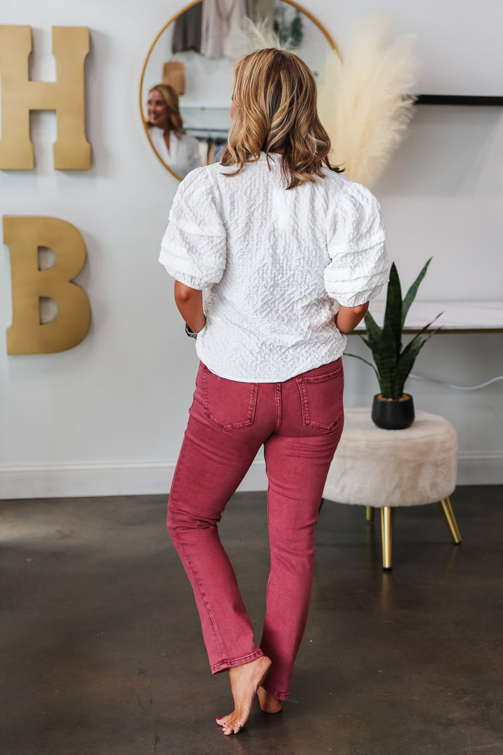 A woman standing in a shop wearing a white shirt and ruby colored jeans with a medium rise and straight leg. She is rear facing.