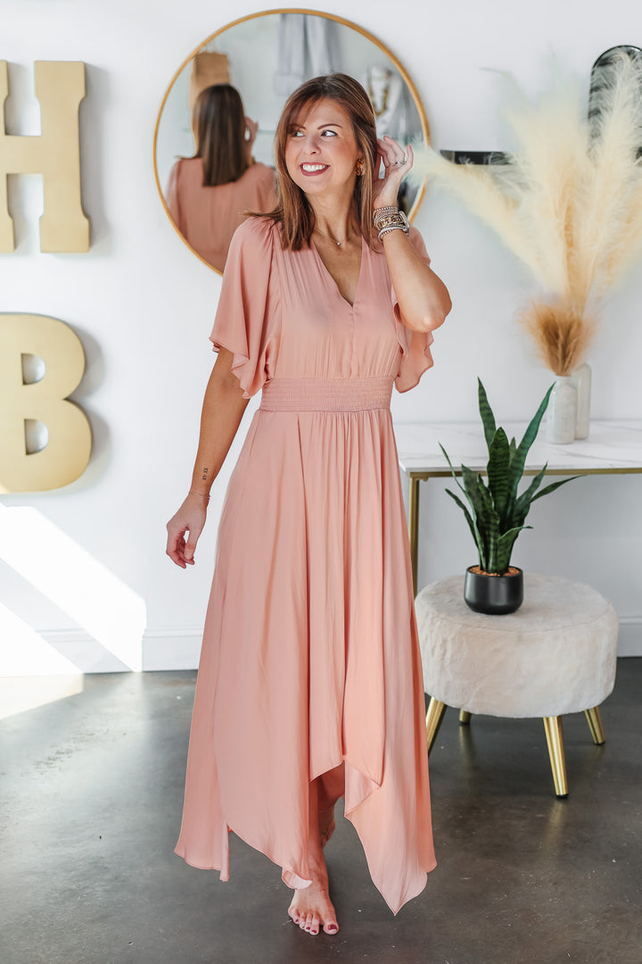 A brunette woman standing in a shop wearing a peach colored dress with asymmetrical hemline, smocked waist and flowy sleeves.