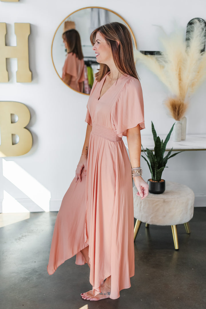 A brunette woman standing in a shop wearing a peach colored dress with asymmetrical hemline, smocked waist and flowy sleeves.