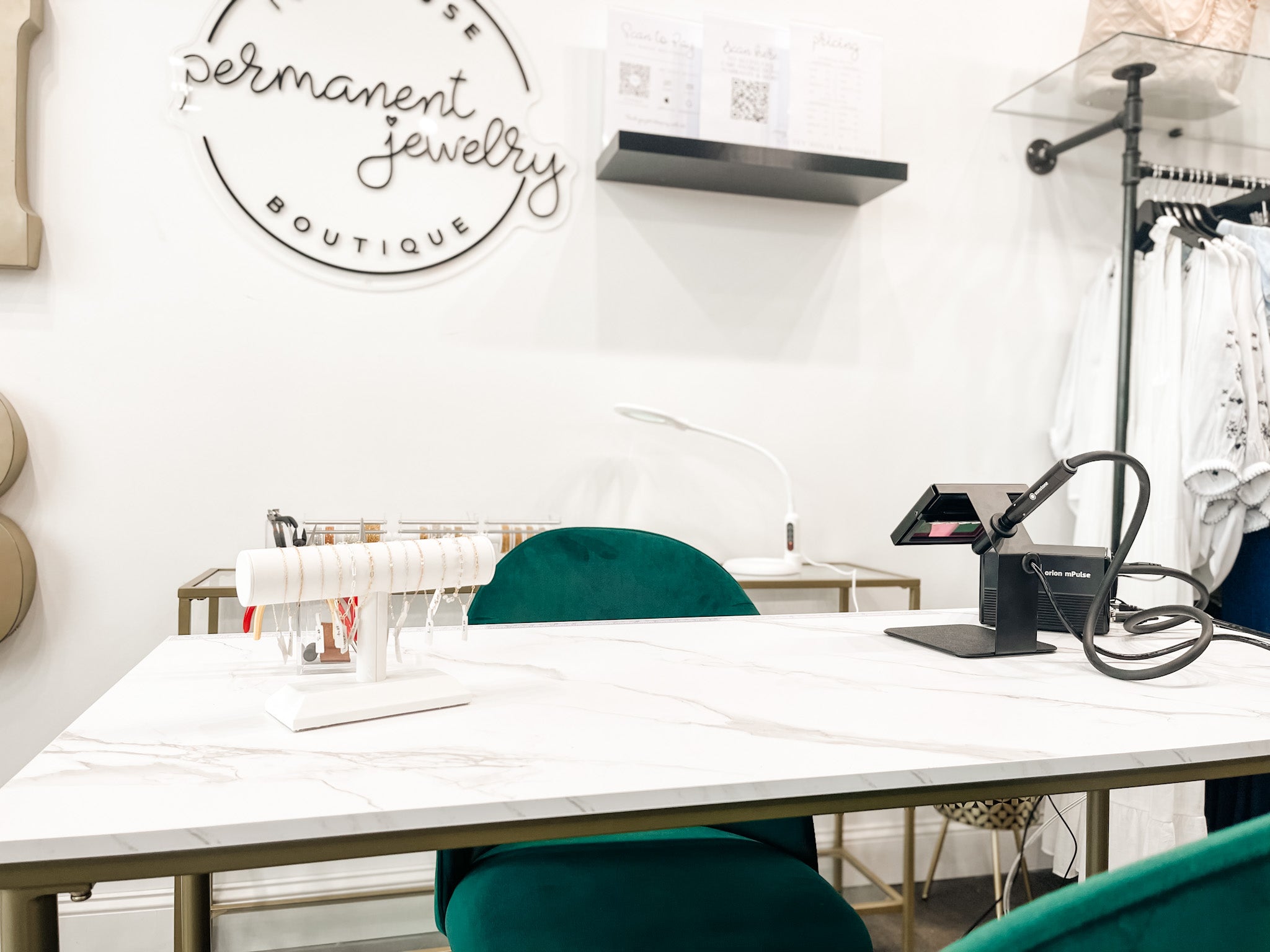 Photo of the permanent jewelry section in Ivy House Boutique. It features a white and gold table, two green chairs, a welding machine, and white bracelet bar displaying various chains. The wall behind it is white.