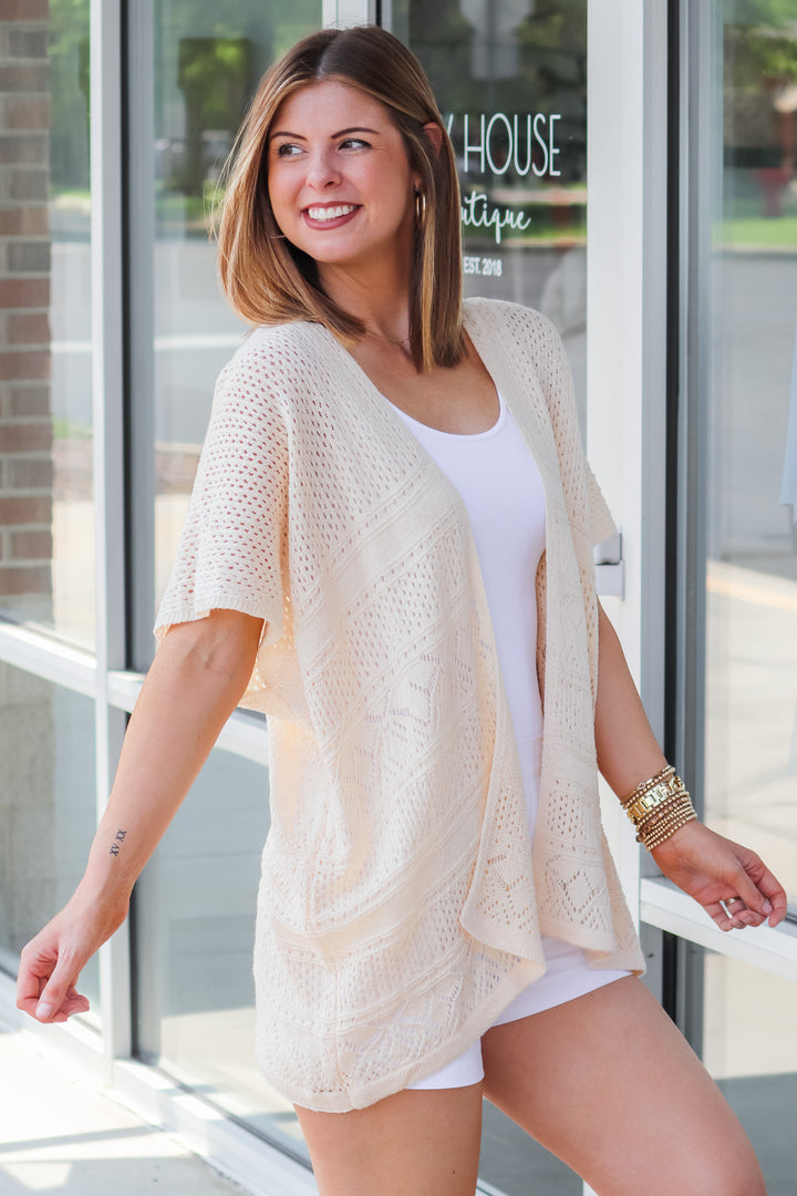 A brunette woman wearing a cream colored short sleeve crochet sweater and white tank. She is standing in front of a shop.