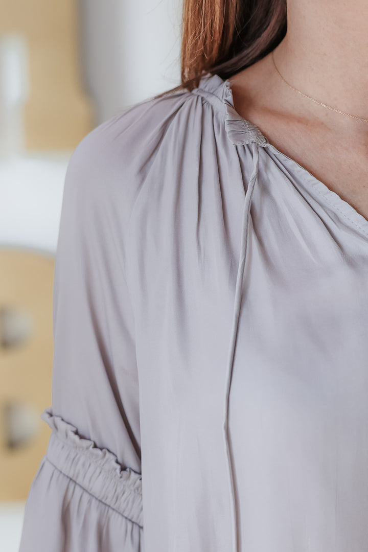 The close up view of the collar of a satin gray top with tie collar and sleeve detail.