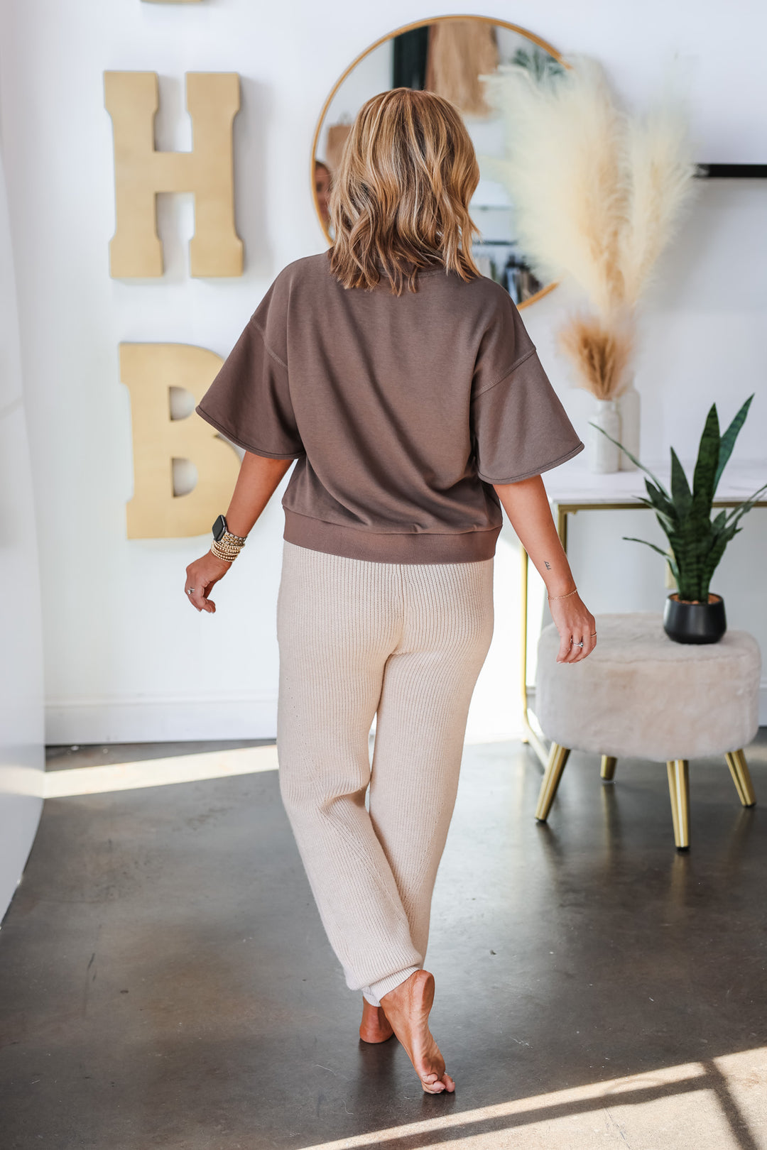 A woman standing in a shop wearing a brown short sleeve sweatshirt and taupe knit joggers with oversized front pockets. She is rear facing.