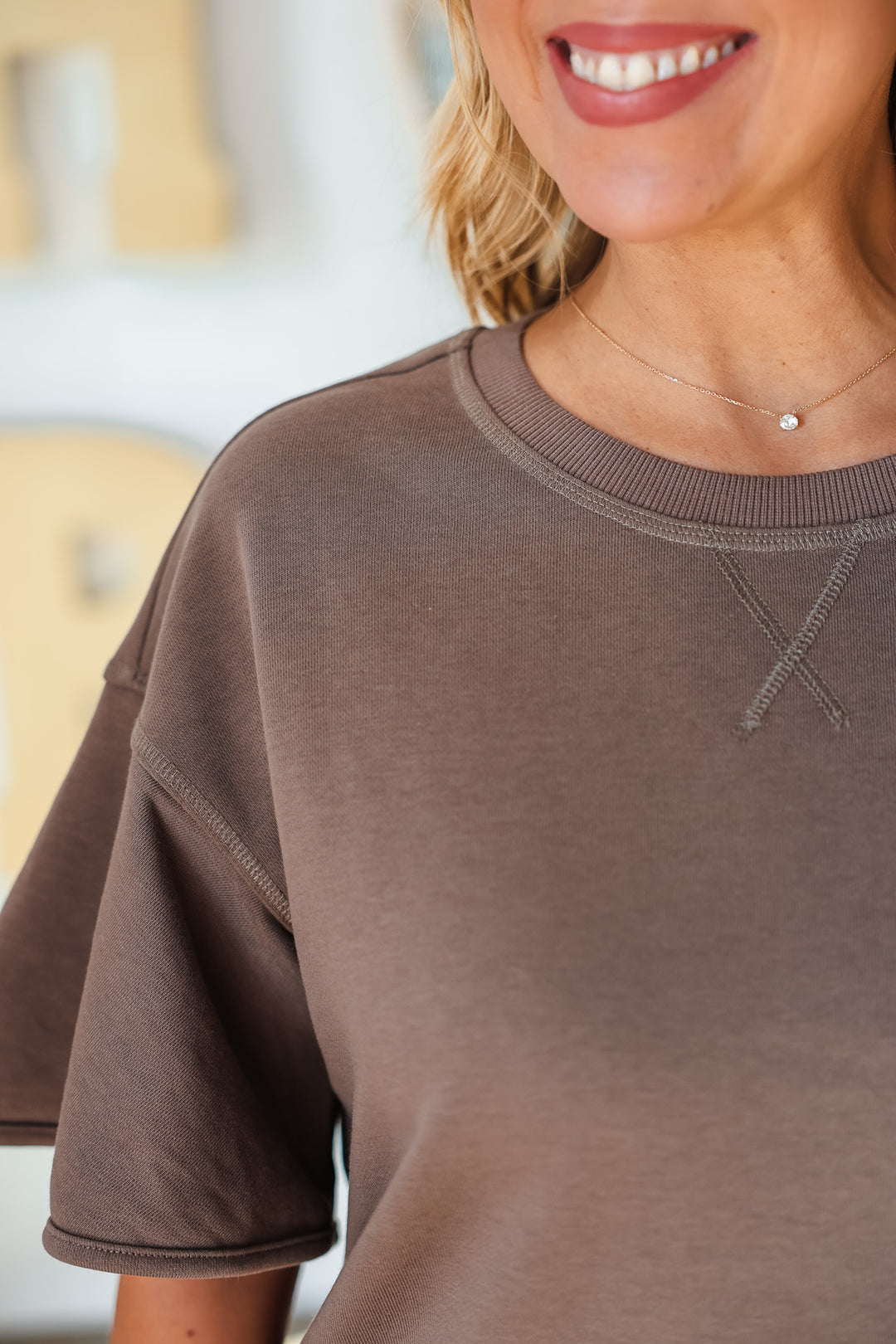 A closeup of a blonde woman standing in a shop wearing a brown sweatshirt with a wide sleeve.