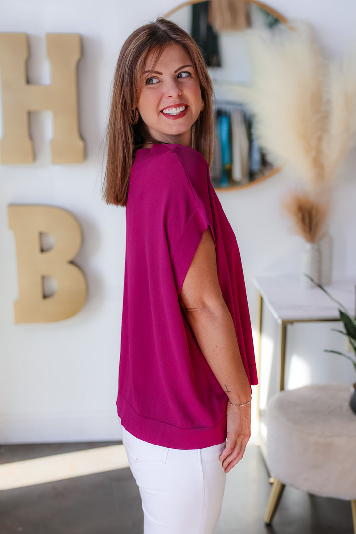 A brunette woman standing in a shop wearing a purple oversized boatneck top and white jeans. 