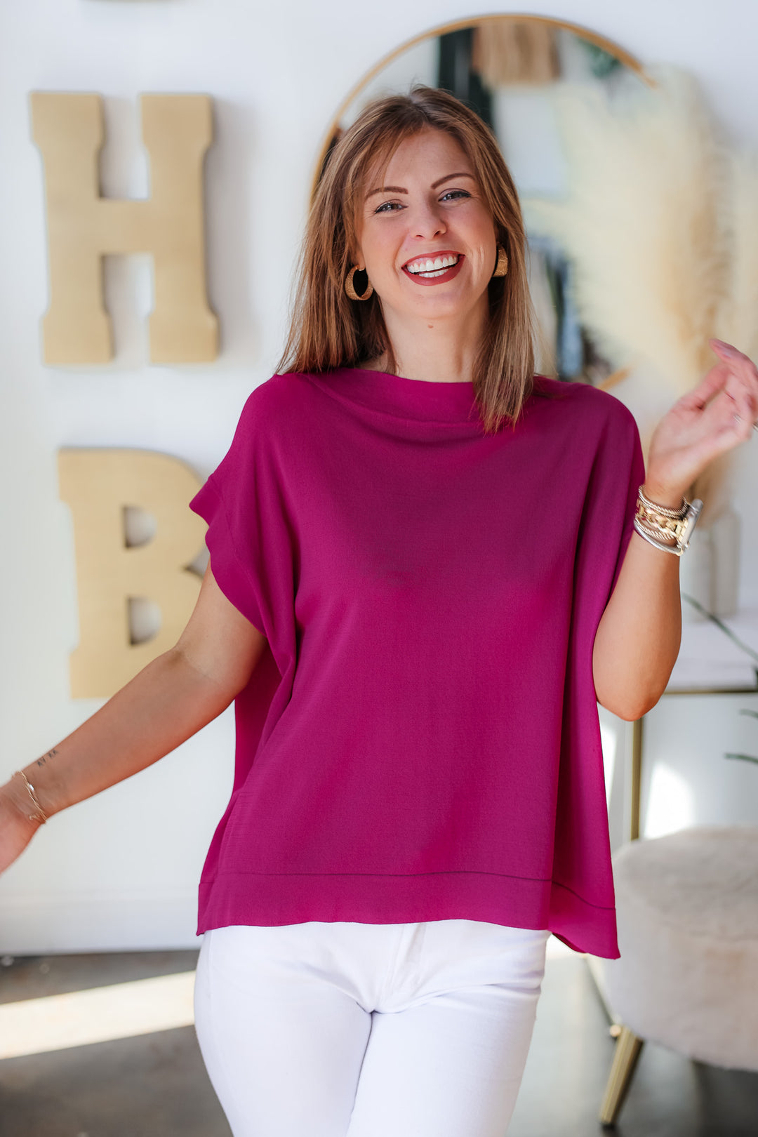 A brunette woman standing in a shop wearing a purple oversized boatneck top and white jeans.