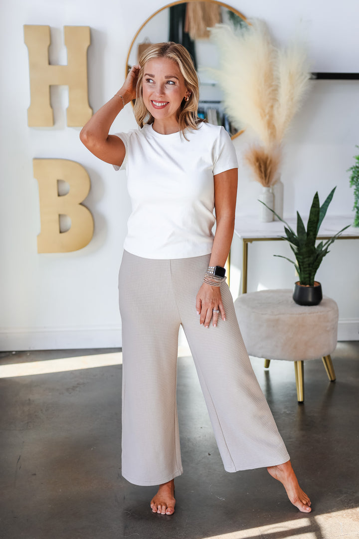 A blonde woman standing in a shop wearing a white tee shirt and wide leg ribbed cream pants.