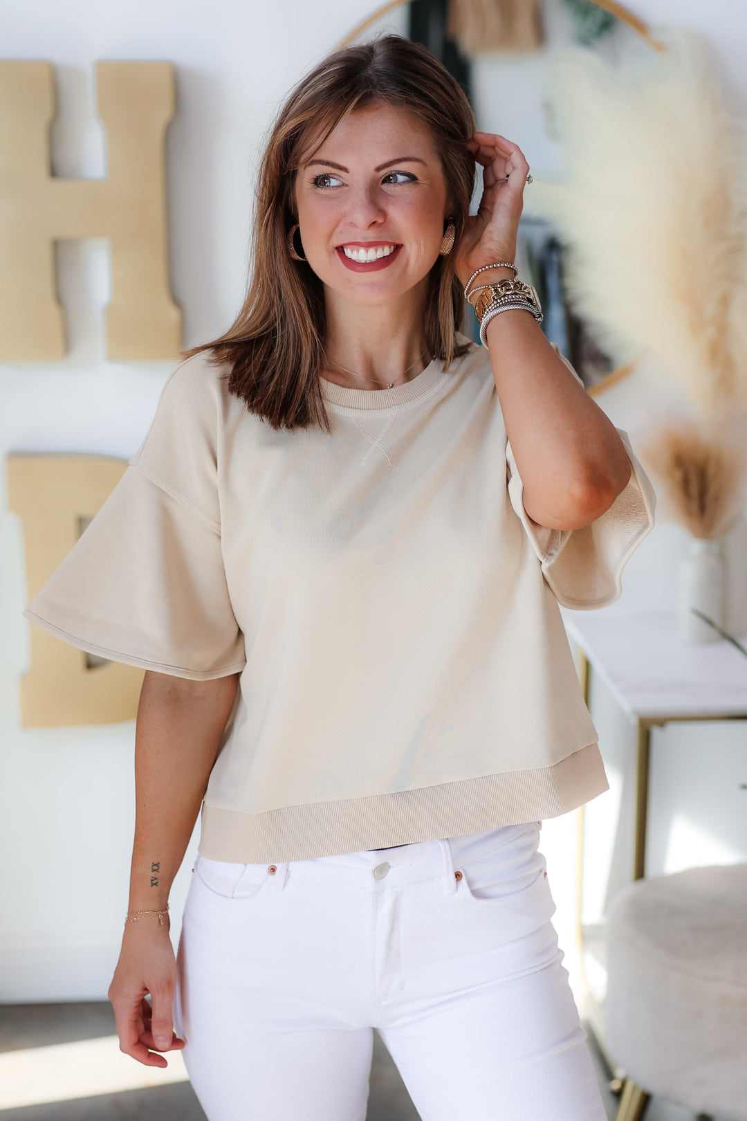 A brunette woman standing in a shop wearing a tan colored short sleeve shirt with wide sleeve and white jeans.
