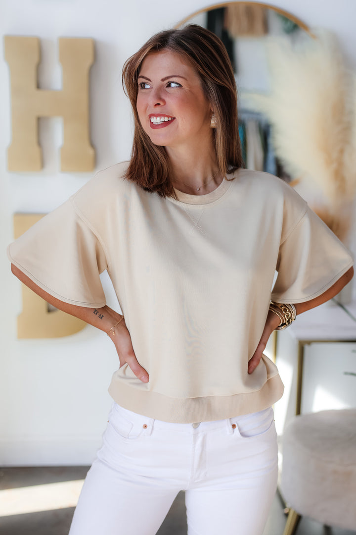 A brunette woman standing in a shop wearing a tan colored short sleeve shirt with wide sleeve and white jeans.