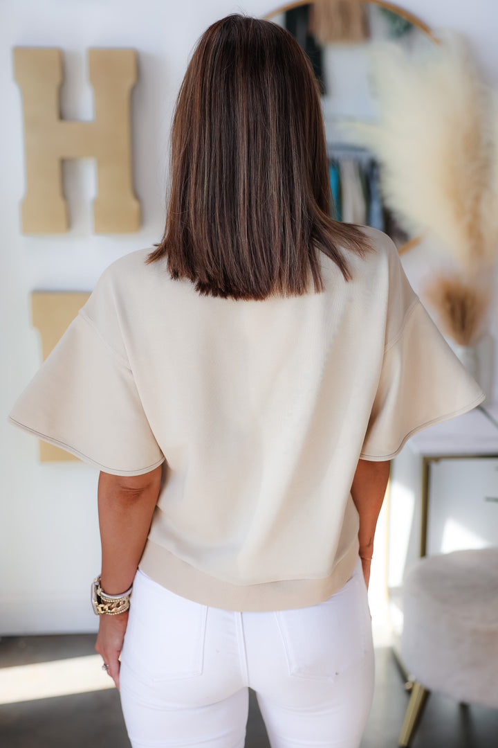 A brunette woman standing in a shop wearing a tan colored short sleeve shirt with wide sleeve and white jeans. She is rear facing.