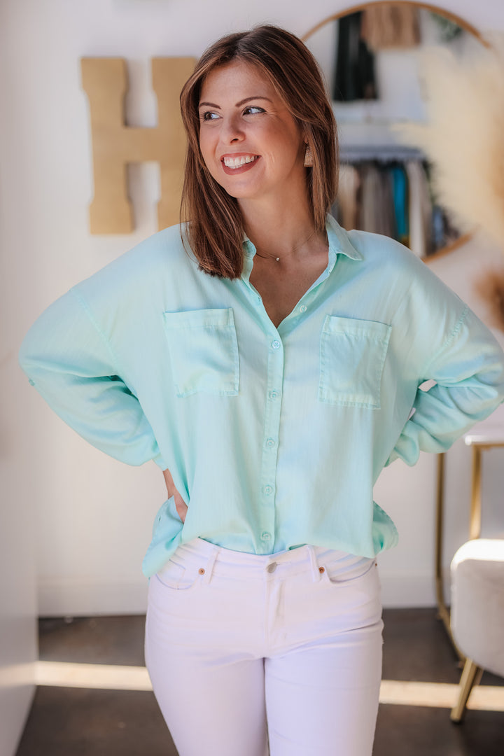 A brunette woman standing in a shop wearing a mint green button down top with white jeans.