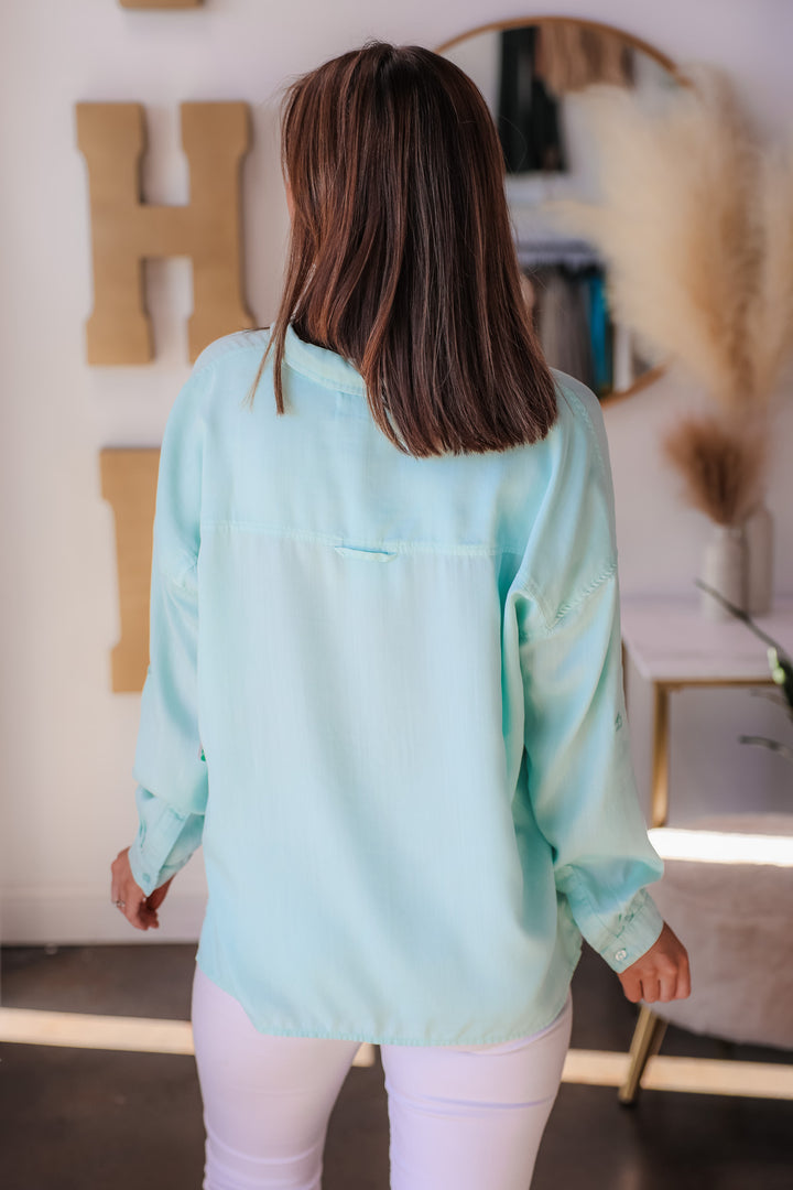 A brunette woman standing in a shop wearing a mint green button down top with white jeans. She is rear facing.