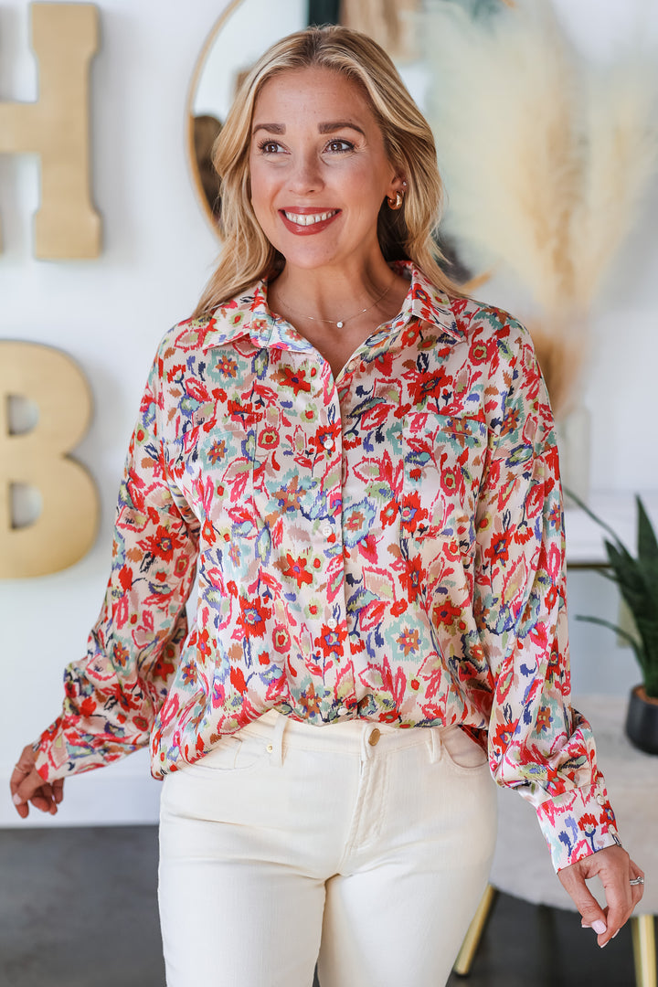 A blonde woman standing in a shop wearing a floral printed button down top. The colors in the top are pink, red, blue, green and cream. She is wearing it with ivory colored jeans.