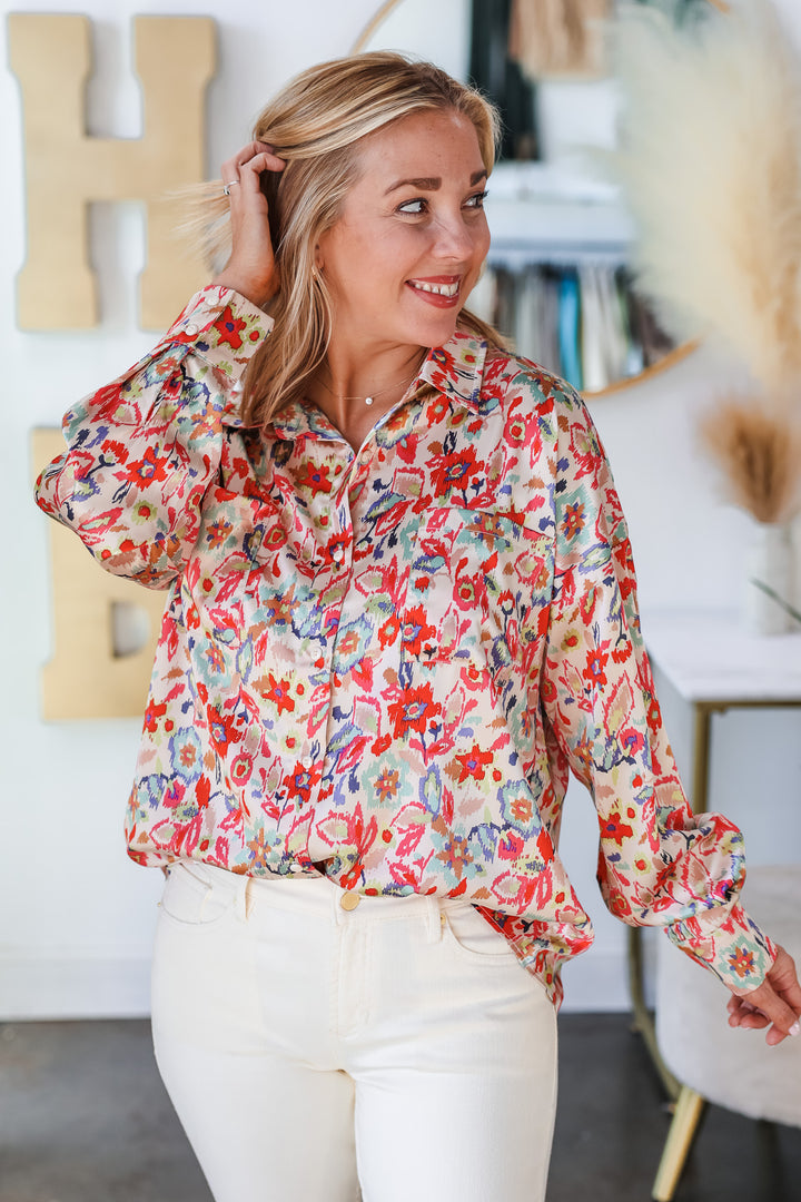 A blonde woman standing in a shop wearing a floral printed button down top. The colors in the top are pink, red, blue, green and cream. She is wearing it with ivory colored jeans.