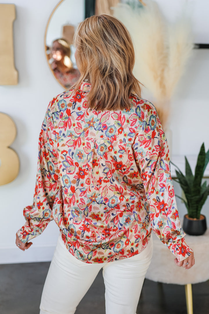 A blonde woman standing in a shop wearing a floral printed button down top. The colors in the top are pink, red, blue, green and cream. She is wearing it with ivory colored jeans. She is rear facing.