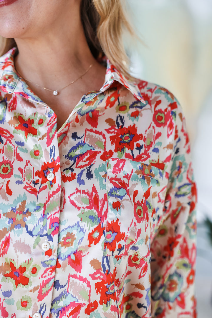 A closeup of the shoulder of a woman wearing a satin printed button down top with pocket. The colors in the top are red, pink green, blue and cream.