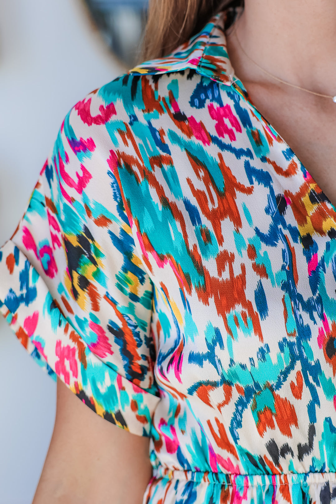 A closeup of the shoulder of a woman wearing a short sleeve abstract print dress with colors of teal, blue, green, pink, yellow, orange and white.