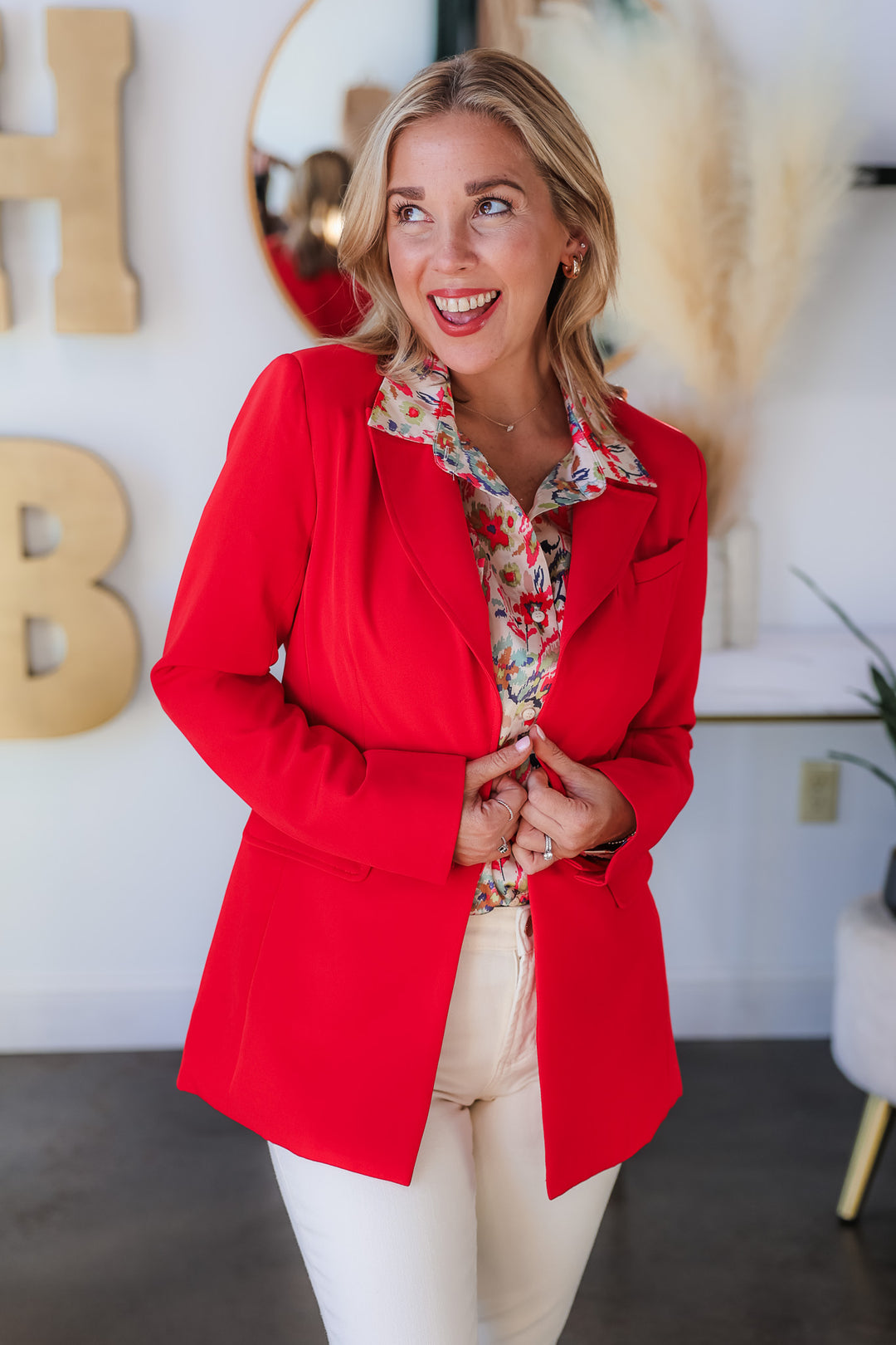 A blonde woman standing in a shop wearing a printed blouse, red blazer and ivory colored blue jeans.