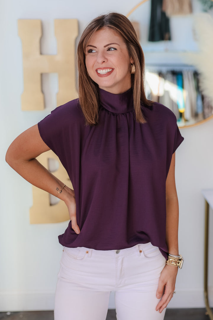 A brunette woman standing in a shop wearing a purple high neck short sleeve blouse with white jeans.