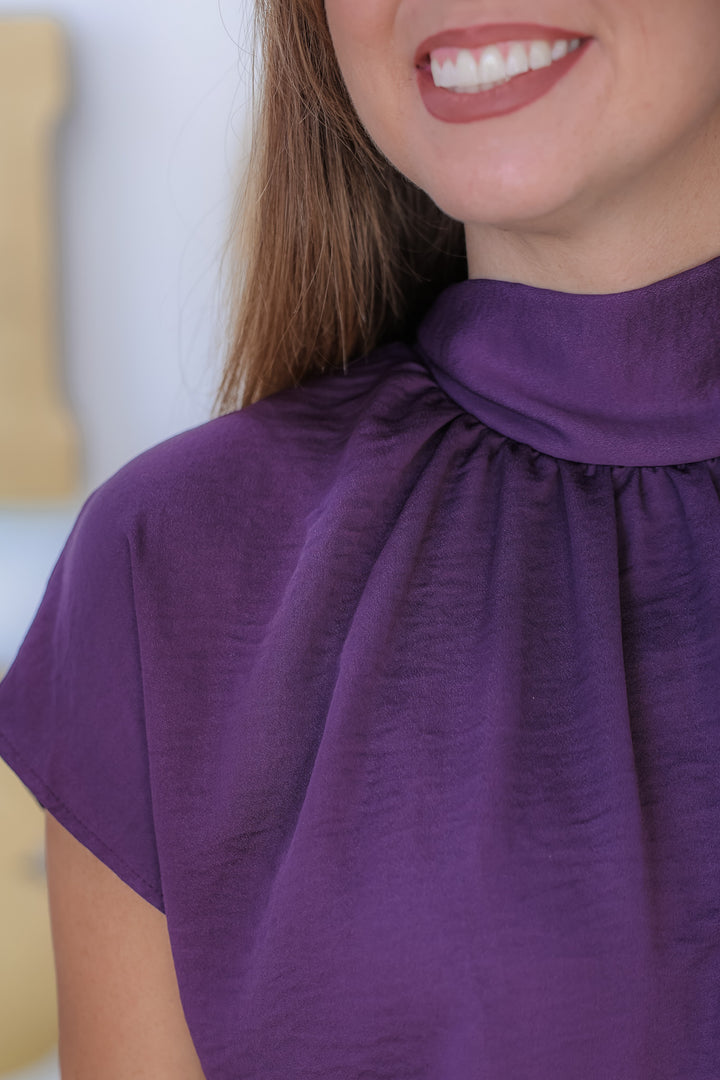 A closeup of a woman wearing a purple high neck blouse with short sleeves.