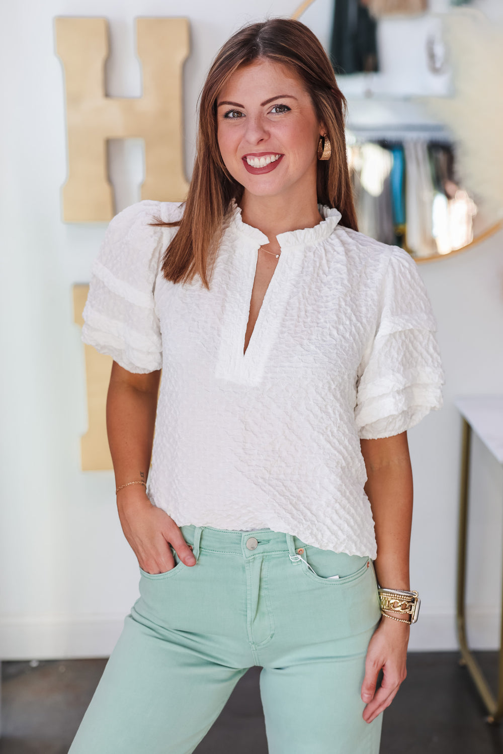 A brunette woman standing in front of a shop wearing a textured short sleeve top with v neck and ruffle collar. She is wearing it with mint colored jeans.