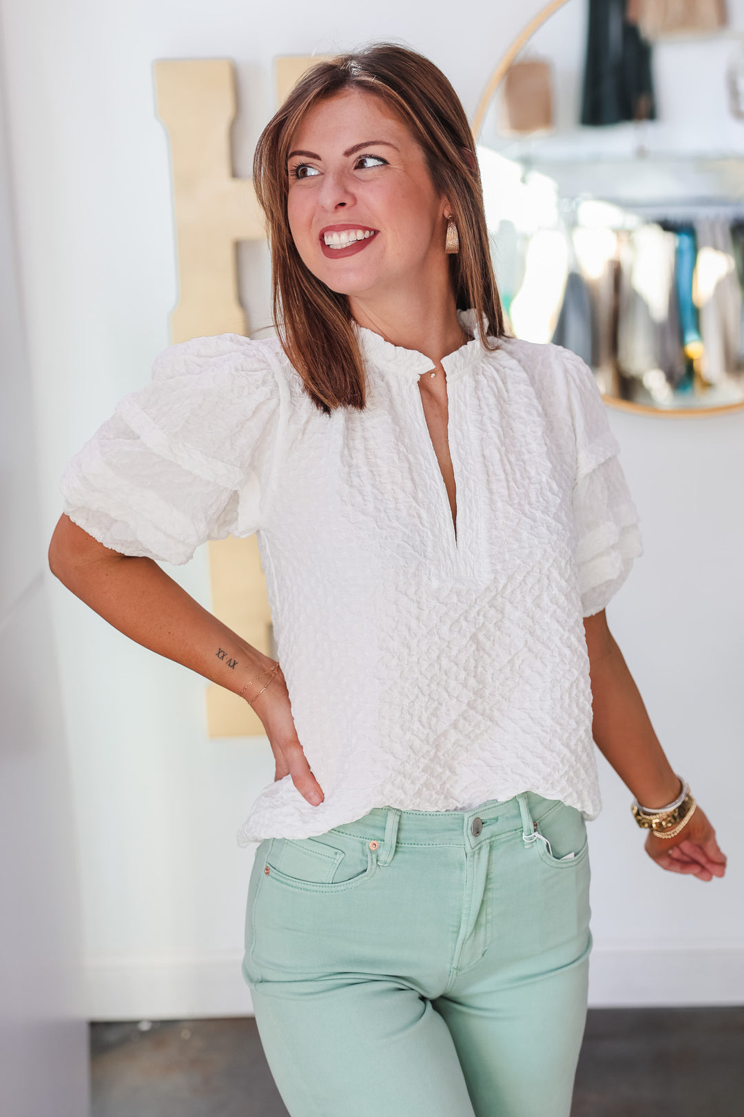 A brunette woman standing in front of a shop wearing a textured short sleeve top with v neck and ruffle collar. She is wearing it with mint colored jeans.