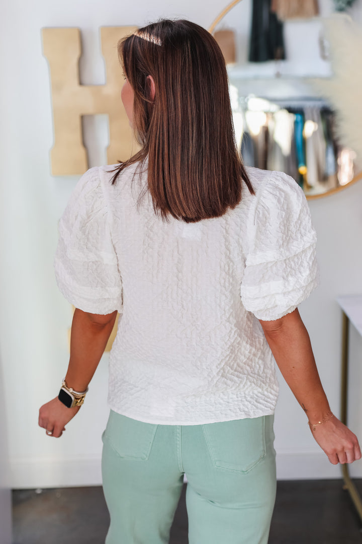 A brunette woman standing in front of a shop wearing a textured short sleeve top with v neck and ruffle collar. She is wearing it with mint colored jeans. She is rear facing.