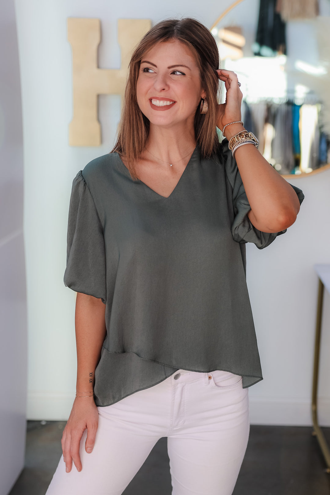 A brunette woman standing in a shop wearing an ash green, short bubble sleeve, v neck top with white jeans.