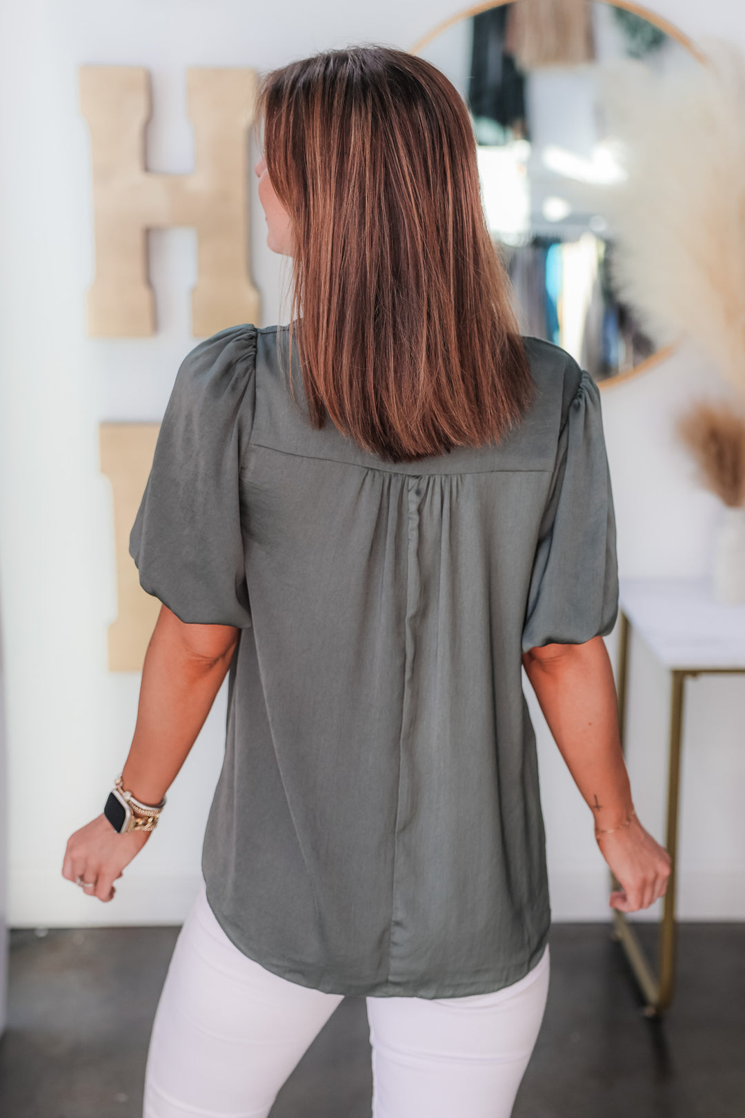 A brunette woman standing in a shop wearing an ash green, short bubble sleeve, v neck top with white jeans. She is rear facing.