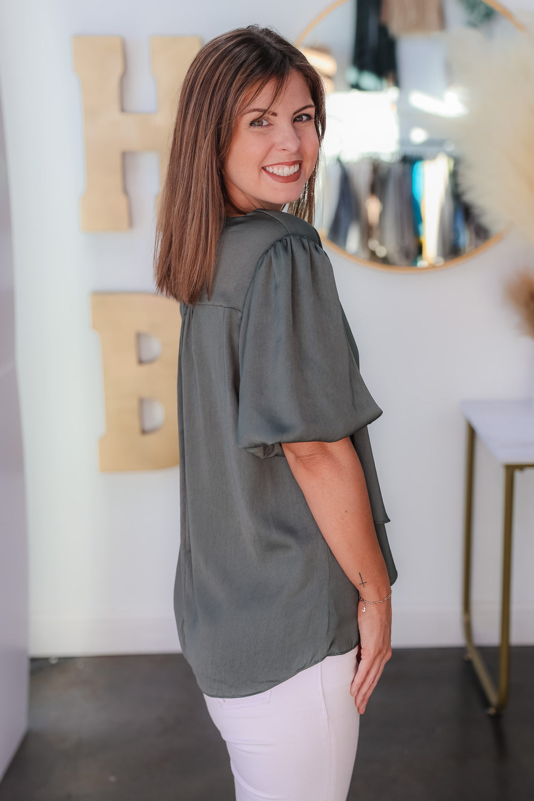 A brunette woman standing in a shop wearing an ash green, short bubble sleeve, v neck top with white jeans.