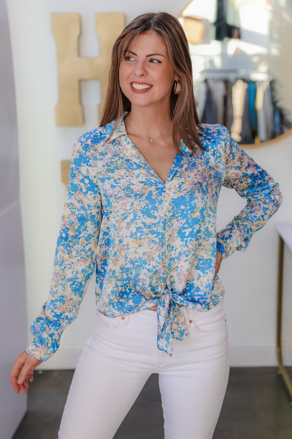 A brunette woman standing in a shop wearing a satin tie front blouse with white jeans. The colors in the top are blue, pink, white, yellow and cream.