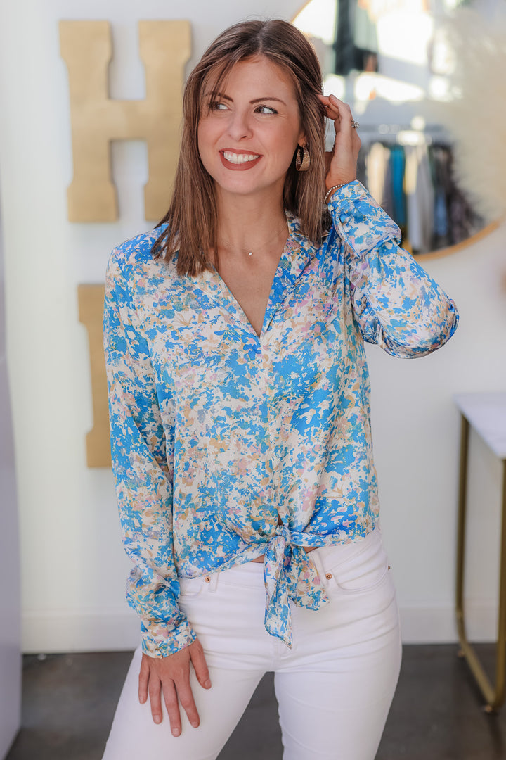 A brunette woman standing in a shop wearing a satin tie front blouse with white jeans. The colors in the top are blue, pink, white, yellow and cream.