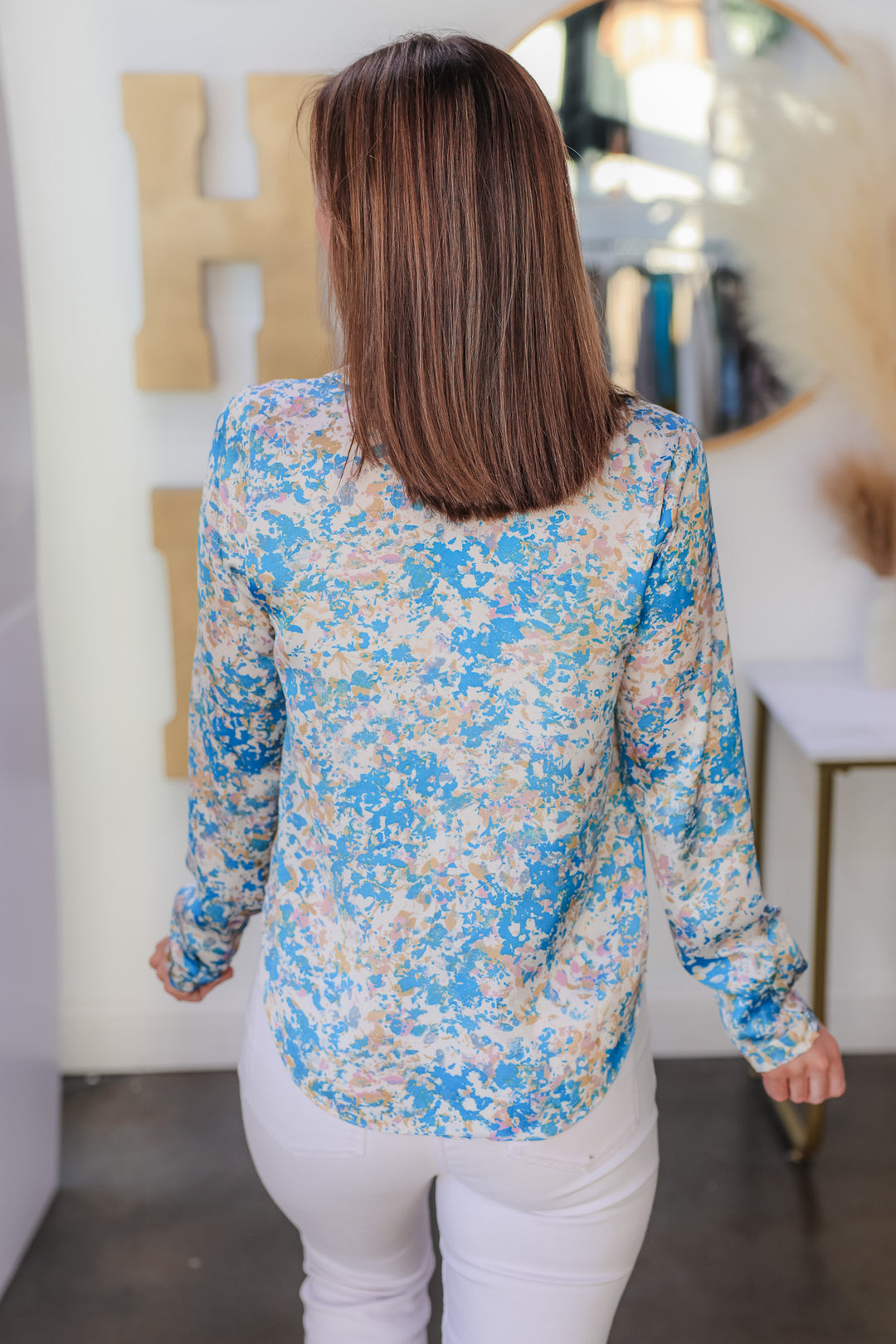 A brunette woman standing in a shop wearing a satin tie front blouse with white jeans. The colors in the top are blue, pink, white, yellow and cream. She is rear facing.