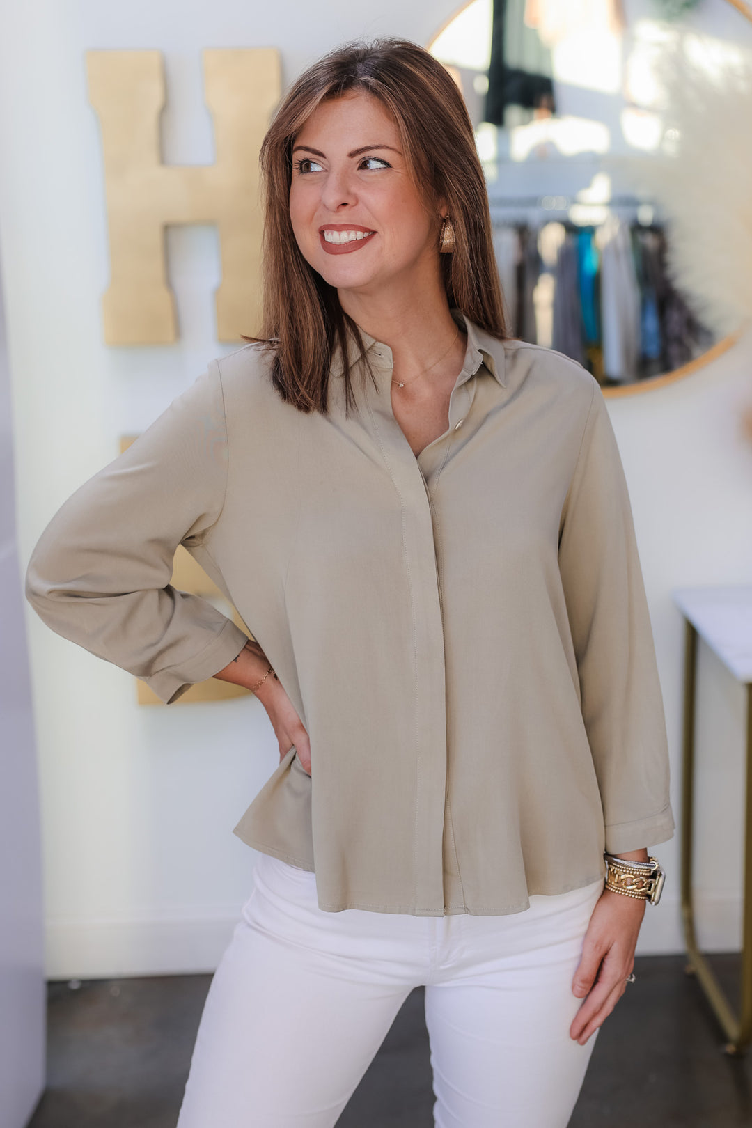 A brunette woman standing in a shop wearing a sage green button down top with white jeans.