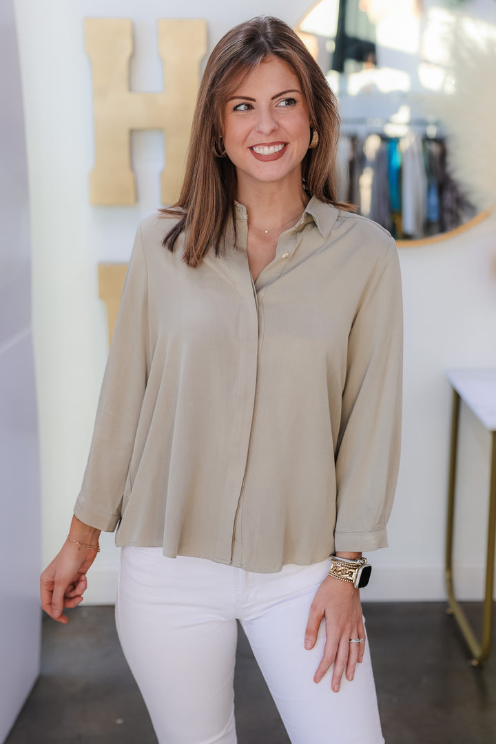 A brunette woman standing in a shop wearing a sage green button down top with white jeans.