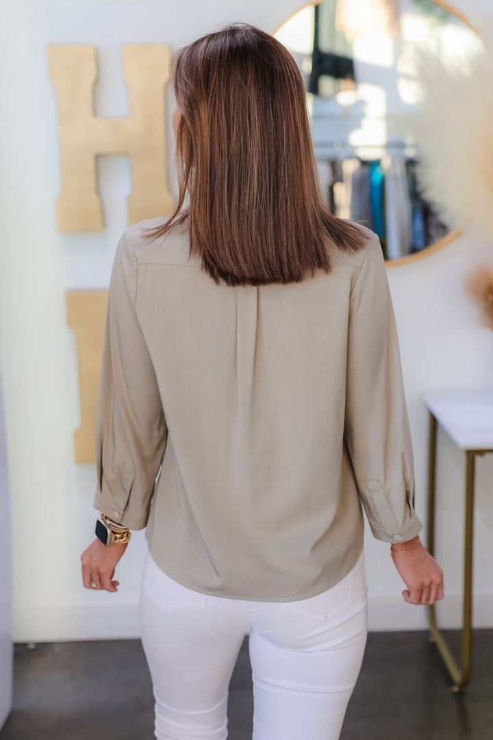 A brunette woman standing in a shop wearing a sage green button down top with white jeans. She is rear facing.
