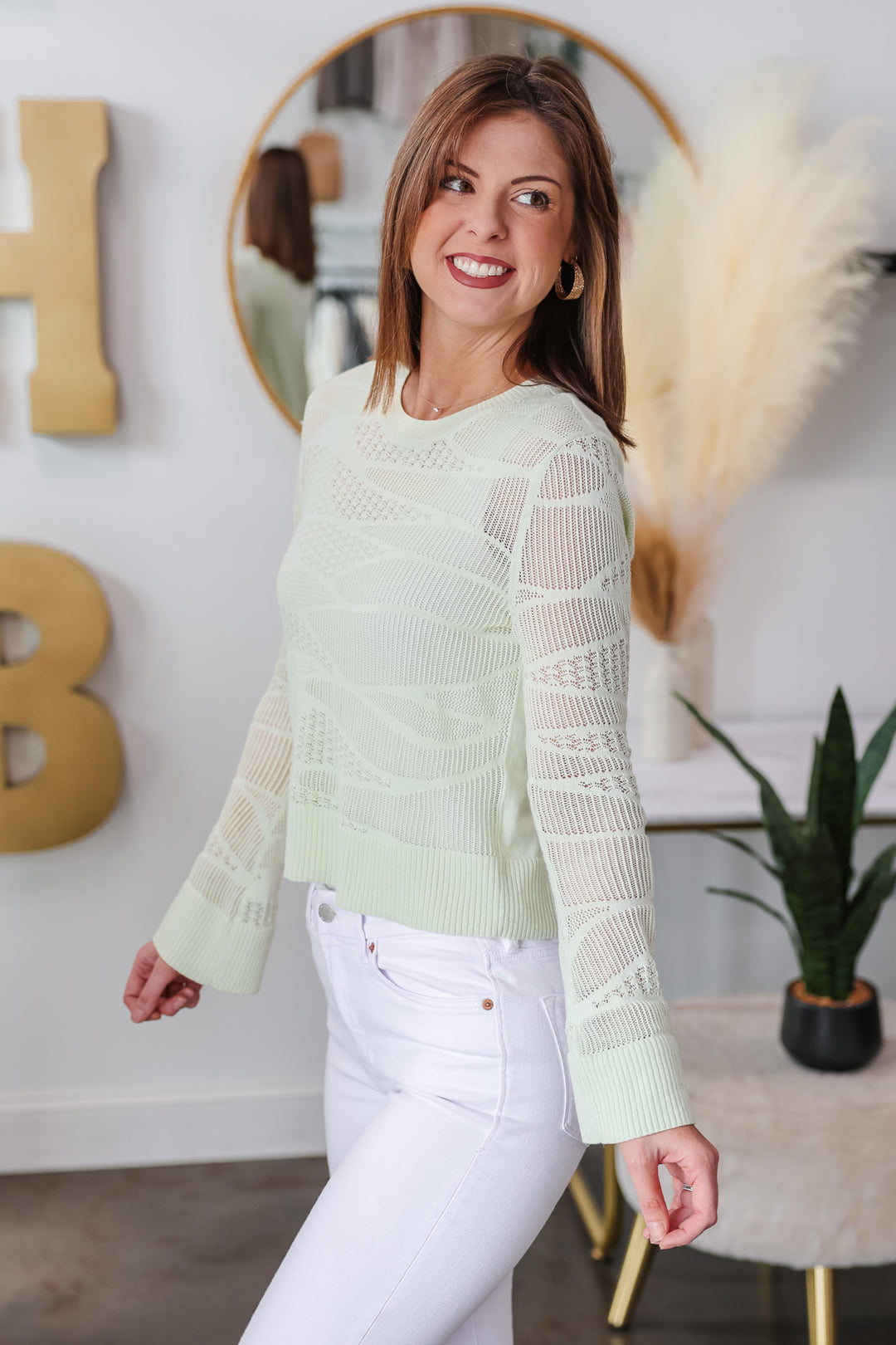 A brunette woman standing in a shop wearing a mint green knit top with white jeans.