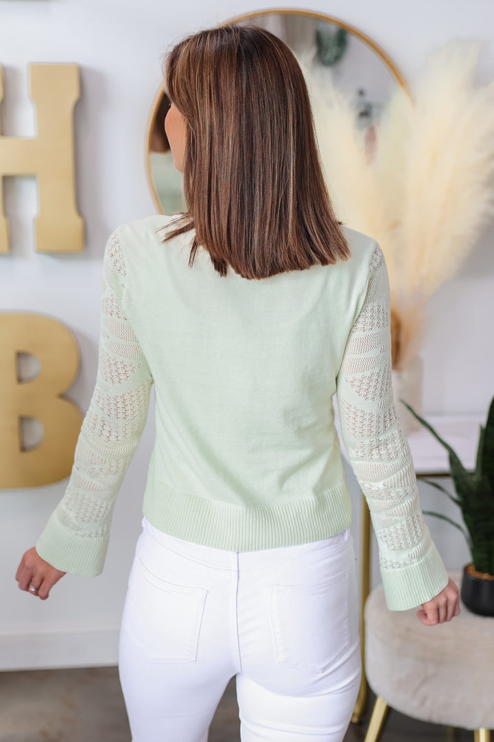 A brunette woman standing in a shop wearing a mint green knit top with white jeans. She is rear facing.