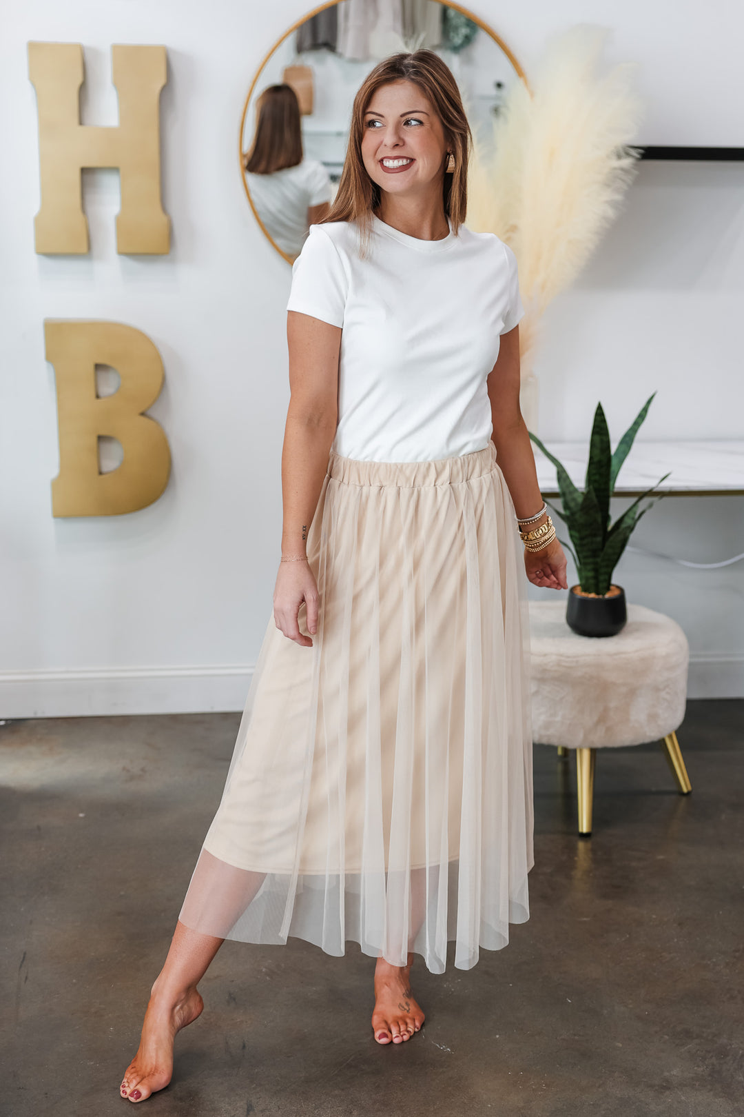 A brunette woman standing in a shop wearing a champagne colored tulle skirt with a basic white tee.