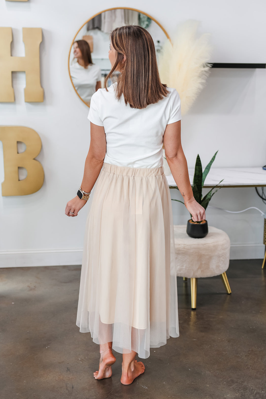 A brunette woman standing in a shop wearing a champagne colored tulle skirt with a basic white tee. She is rear facing.