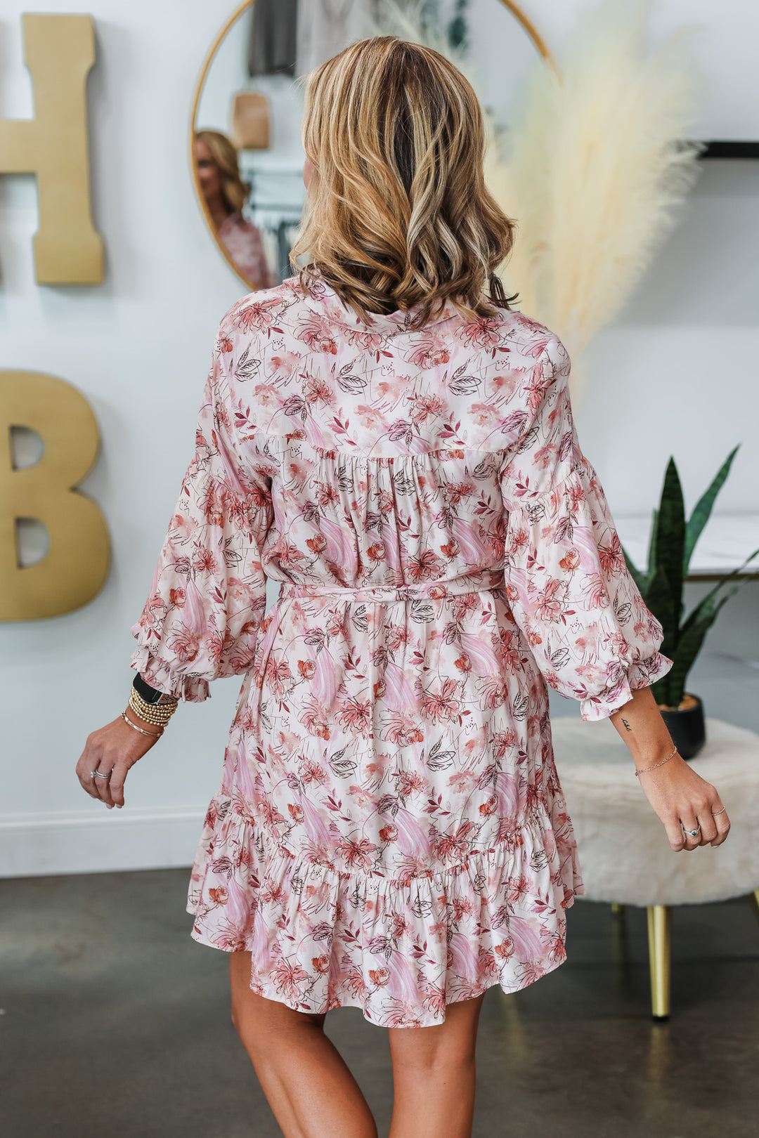A blonde woman standing in a shop wearing a pink bubble sleeve floral shirt dress with adjustable tie belt.