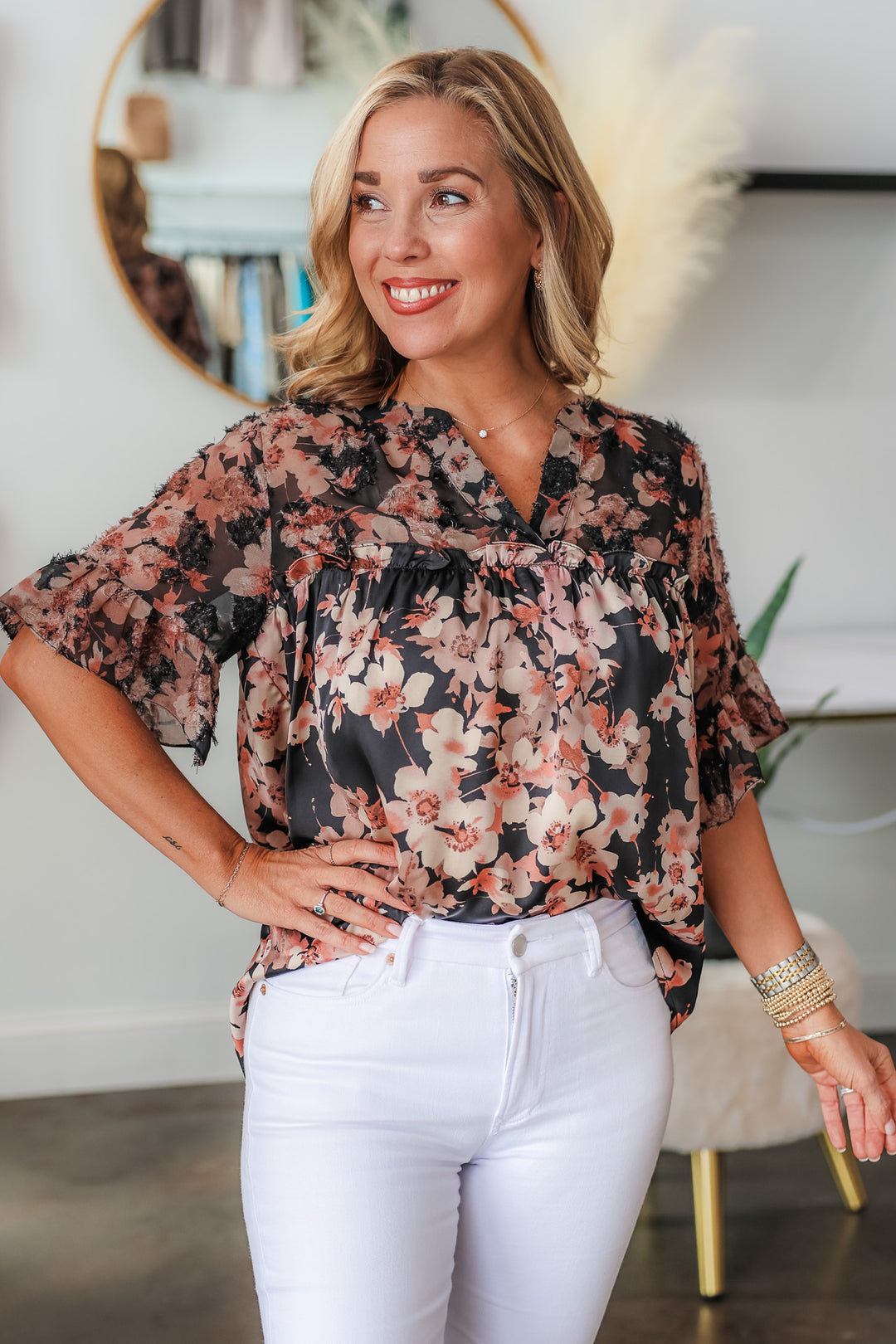 A blonde woman standing in a shop wearing a black and mauve floral print top with ruffle sleeves and a v neck with white jeans.