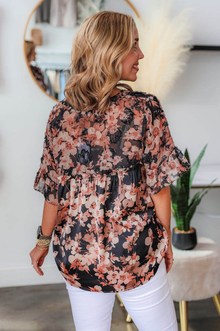 A blonde woman standing in a shop wearing a black and mauve floral print top with ruffle sleeves and a v neck with white jeans. She is rear facing.