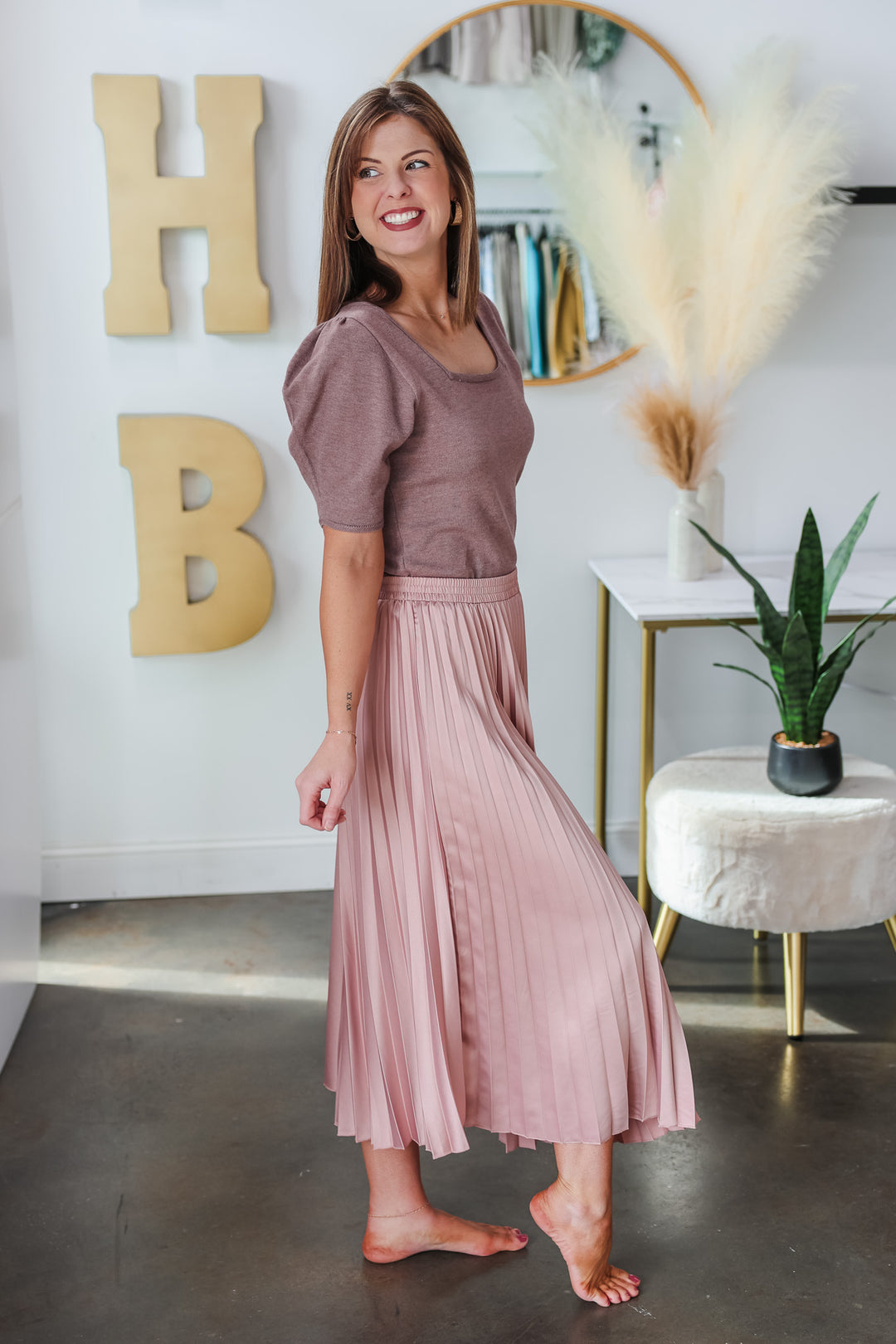 A brunette woman standing in a shop wearing a pink satin pleated midi skirt with elastic waist and taupe top.