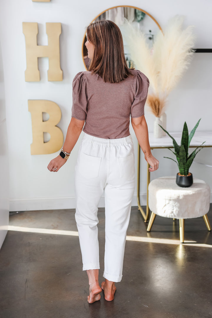 A brunette woman standing in a shop wearing white cotton twill pants with button front and pockets and a taupe sweater. She is rear facing.