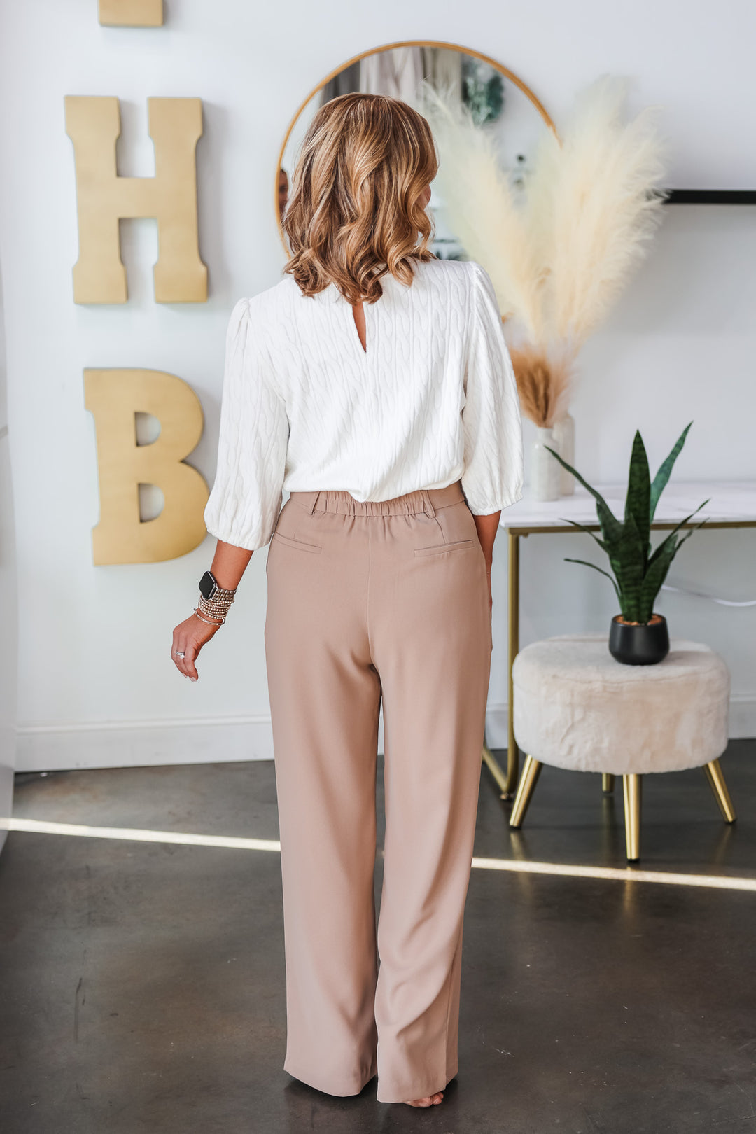 A blonde woman standing in a shop wearing tan dress pants with a wide leg and tab button closure with a white top. She is rear facing.