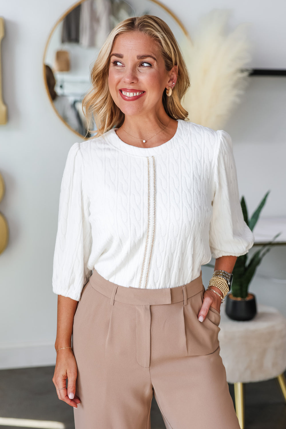 A blonde woman standing in a shop wearing a white cable knit detail top with gold center seam and tan dress pants.
