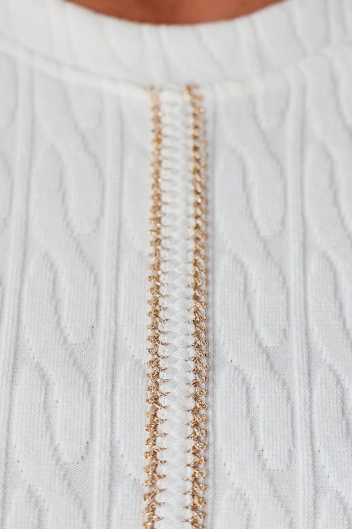 A closeup of the gold trim detail on a white cable knit patterned top.