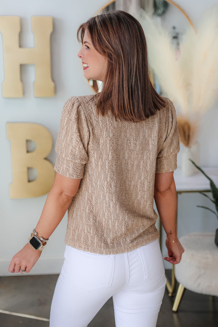 A brunette woman standing in a shop wearing a taupe mock neck textured top and white jeans. She is rear facing.