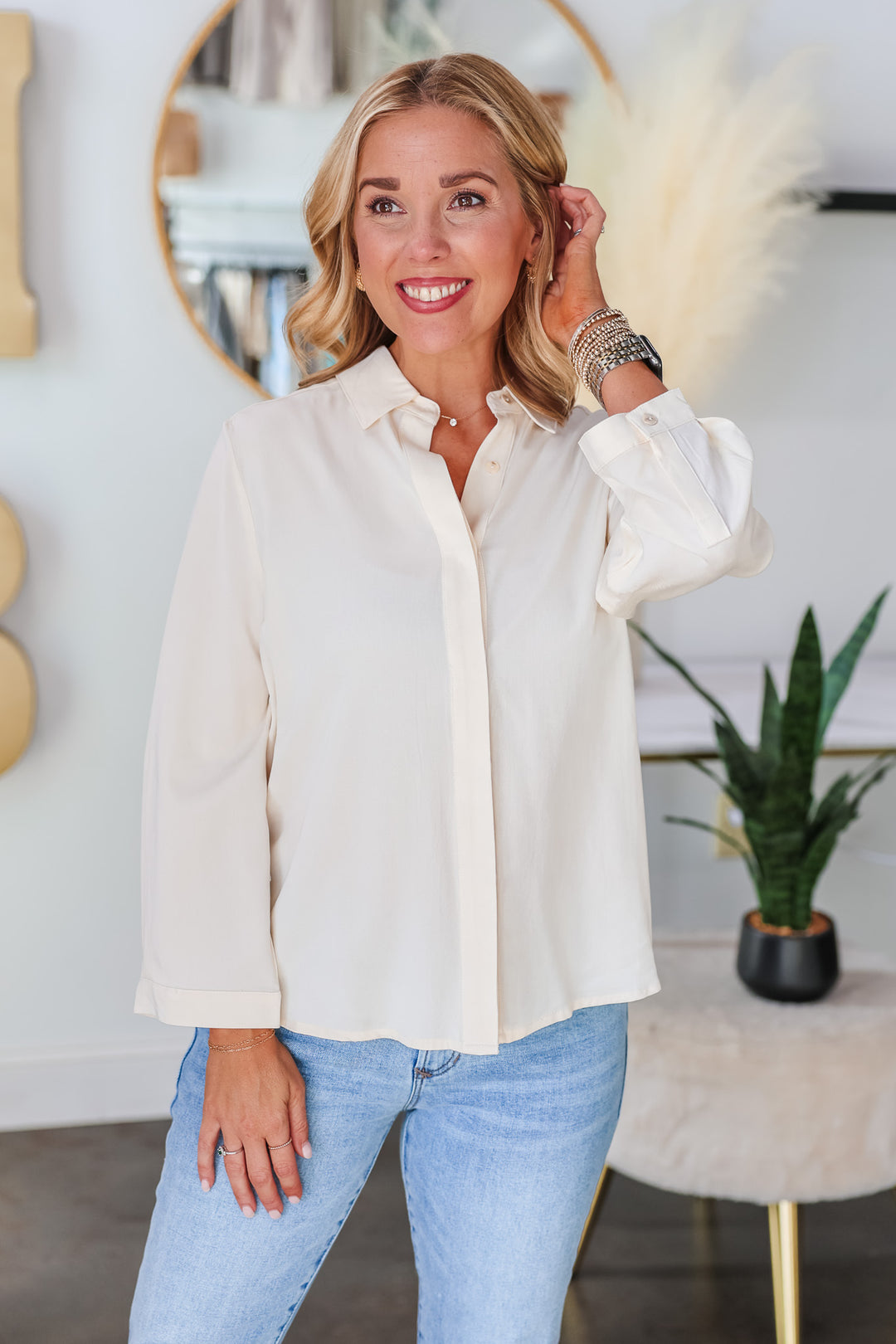 A blonde woman standing in a shop wearing a cream colored button down with blue jeans.